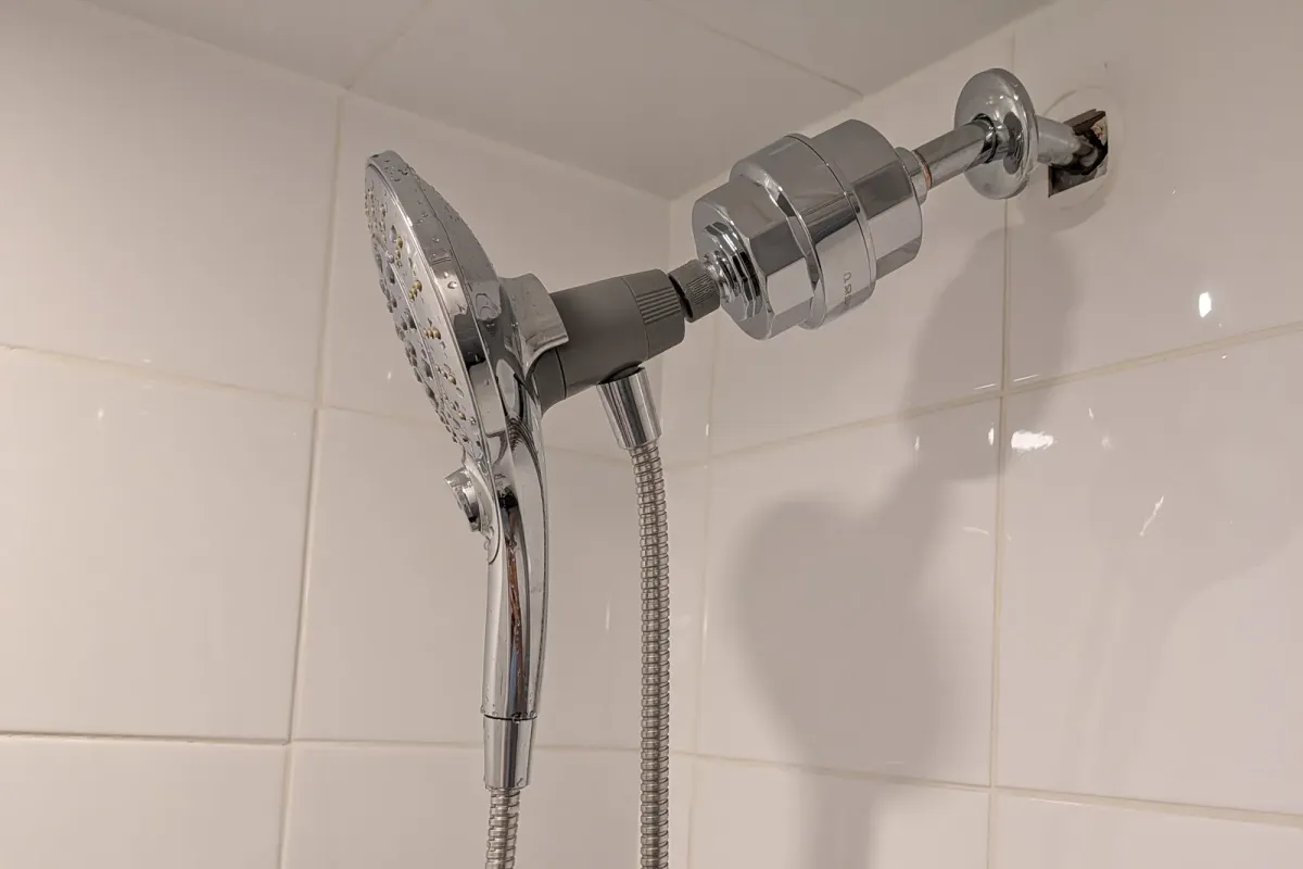 Chrome shower filter and hand shower fitted on a shower arm against white tile