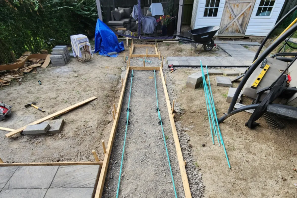 Wooden forms and rebar set on gravel for a backyard walkway, with a shed behind
